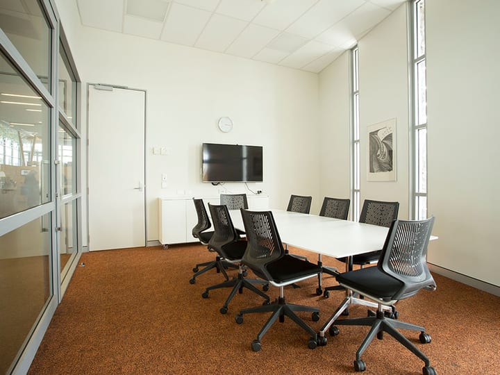 White meeting room with long white table, black chairs, wall-mounted screen and tall windows