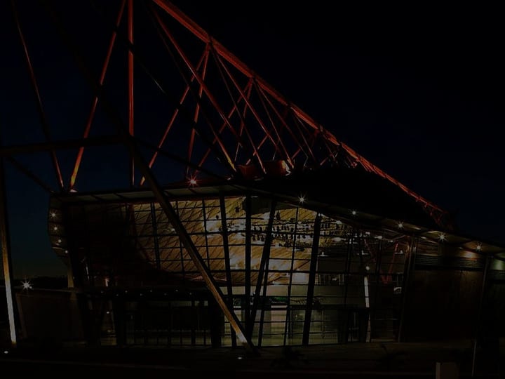 Dramatic event building at night with angled red steel lattice frame, glazed facade and exterior lighting