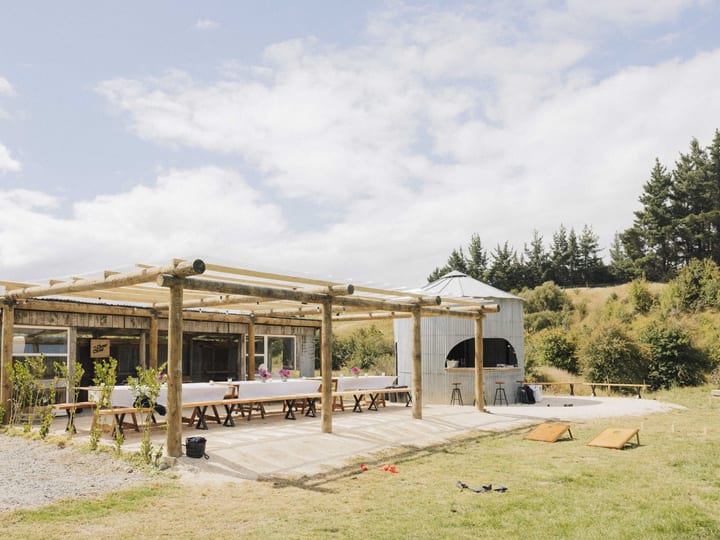 Open timber-frame outdoor shelter with long trestle tables, corrugated iron bar structure beside it, paddock setting