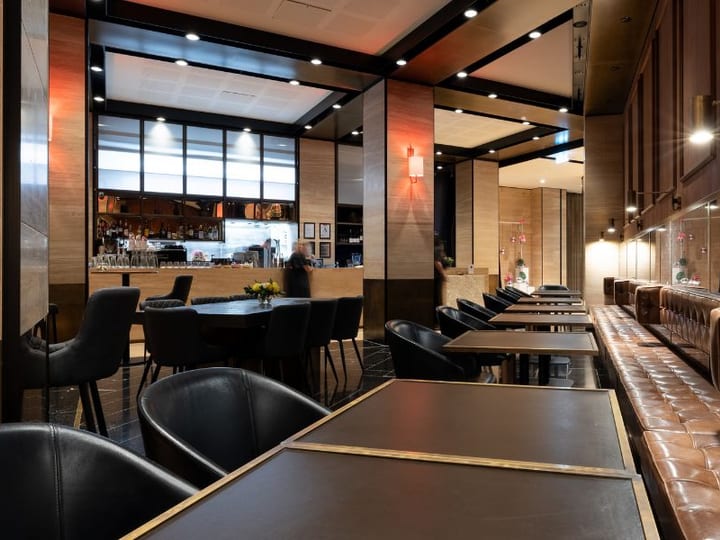 Dark dining room interior with long leather banquette, round tables, warm wall sconces, bar in background