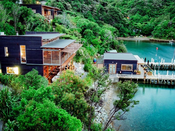 Aerial view of dark-clad lodge buildings set into a steep bush-covered hillside beside a cove