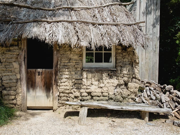 Mud-brick colonial hut with thatched roof, wooden door and bench