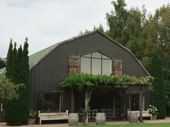 Dark-clad barn venue building viewed across lawn with cypress trees flanking the entrance and wine barrels at the porch