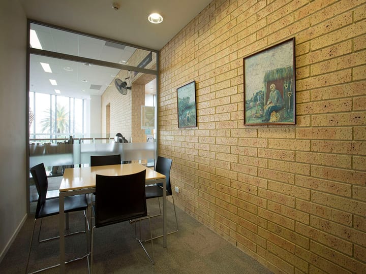 Small meeting room with yellow brick wall, framed artworks, light timber table and dark chairs behind glass partition