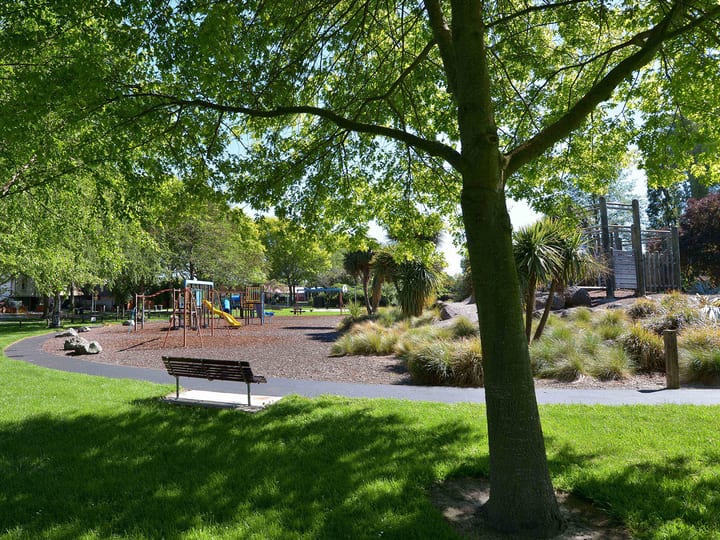 Park with trees, lawn and playground equipment visible, bench in foreground
