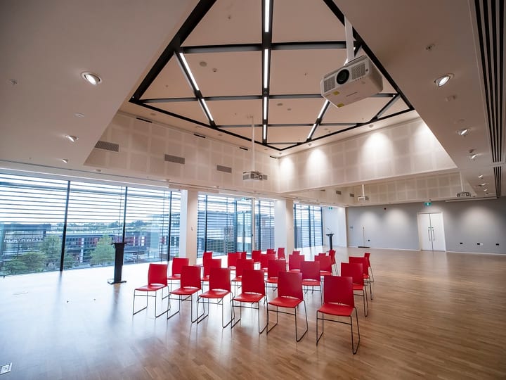 Open hall with timber floor, red chairs in rows, geometric black ceiling frames, and floor-to-ceiling windows