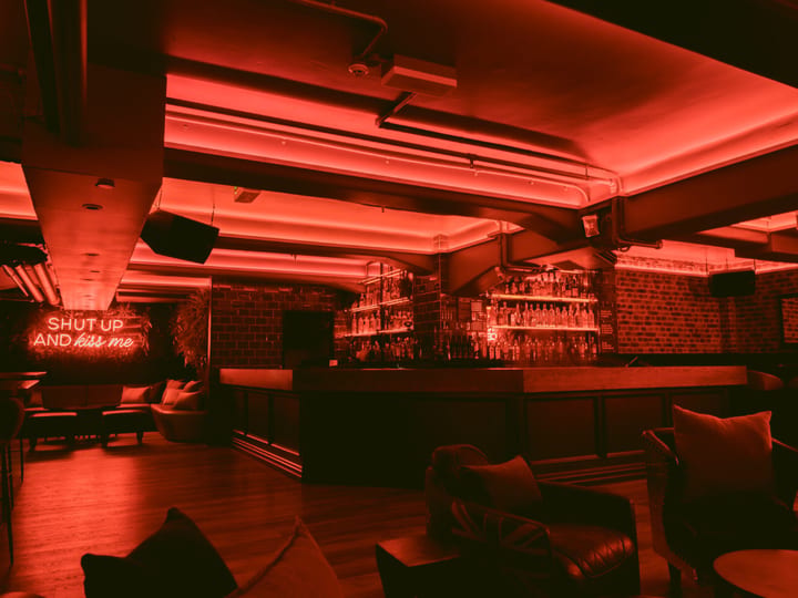 Dark basement bar with red neon lighting, curved bar counter, exposed brick walls, lounge seating