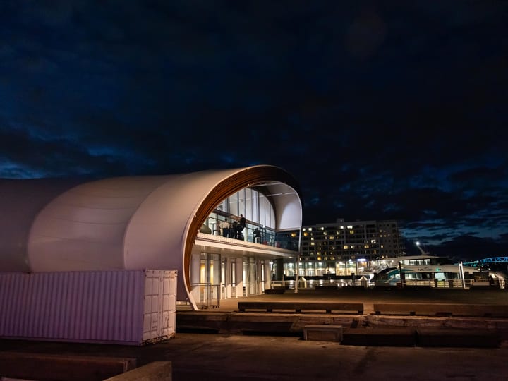 White curved barrel-vault building at night on a waterfront, city lights behind