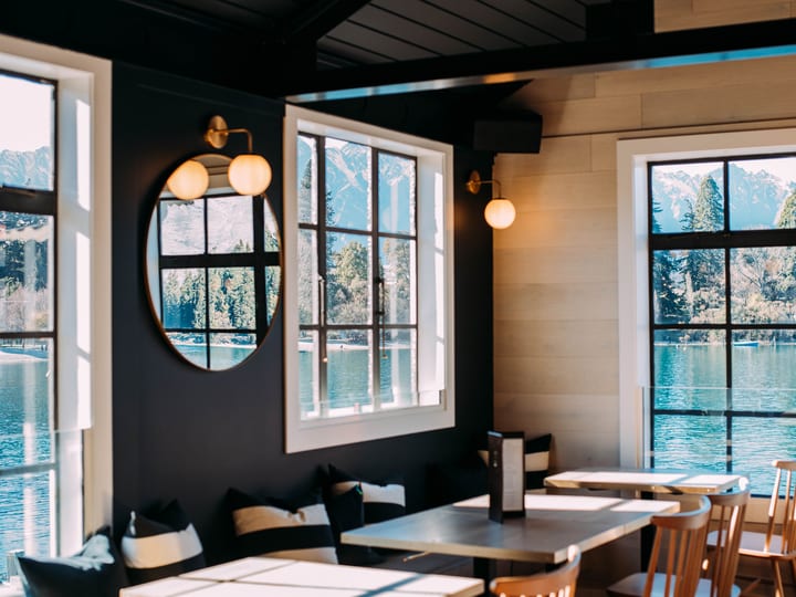 Dark-painted interior with black ceiling beams, round mirror, globe wall lights, and lake and mountain view through white-framed windows