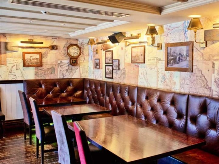 Bar interior with dark timber banquette seating, tufted leather backs, map-print wallpaper, and wall sconce lighting