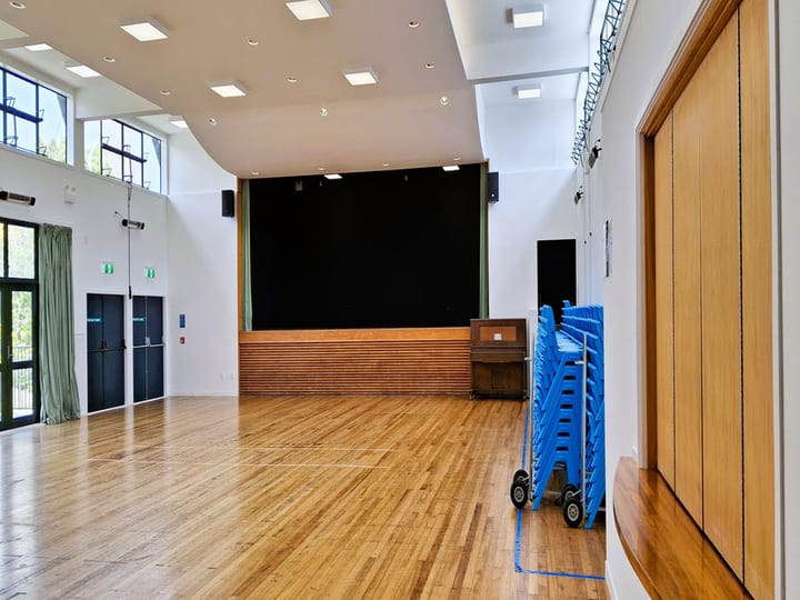 Large hall interior with timber floor, stage platform, stacked chairs and high windows