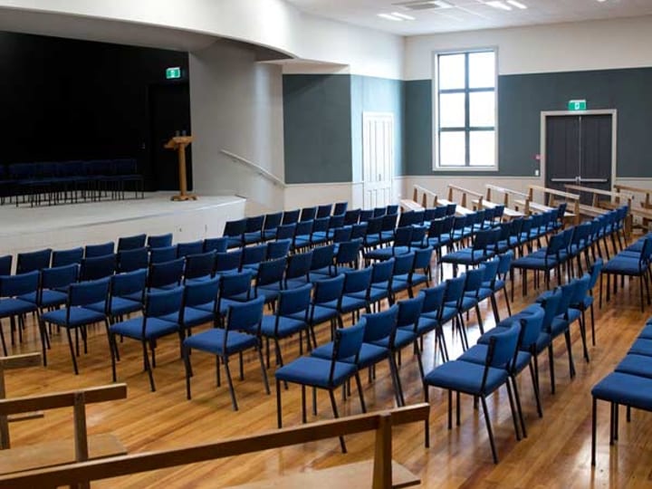 Hall interior with rows of blue chairs facing a raised platform and stage
