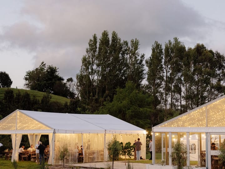 White marquee structures with festoon lights on a hillside at dusk with trees behind