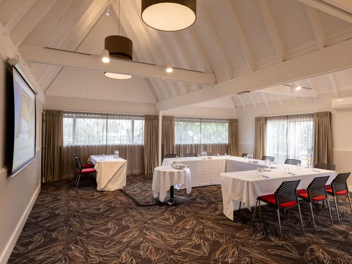 White-plastered function room with vaulted timber-truss ceiling, white tables in U-shape, and large screen