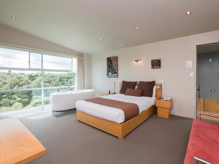 Studio with a king bed and a freestanding bath beside full-height windows over native bush