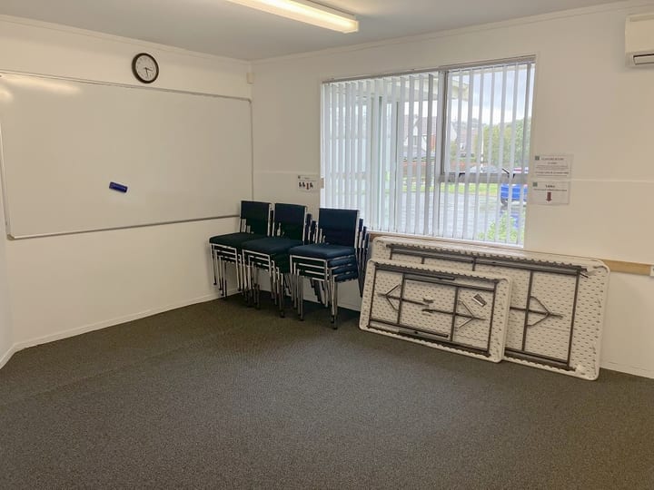 Meeting room with whiteboard, stacked chairs and folded tables, vertical blinds on window