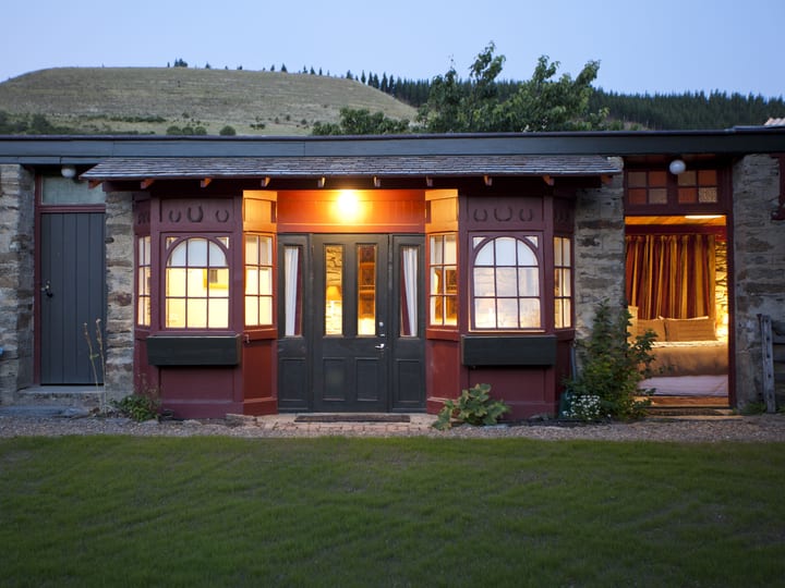 Stone stable building at dusk with lit interiors through arched windows opening onto a lawn