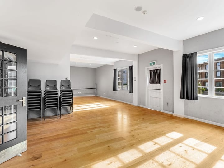 Empty carpeted room with stacked chairs, curtained windows, and kitchenette alcove