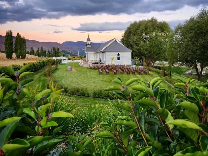 White church with bell tower set on a green lawn with mountains and dramatic sky behind