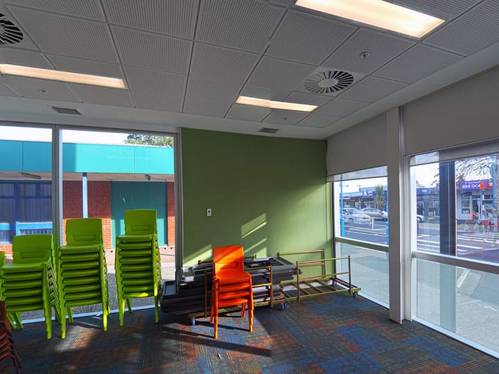 Bright community room with stacked green and orange chairs, large windows to street