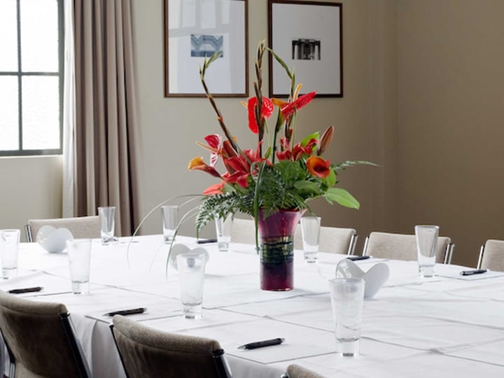 Boardroom table with white linen, floral centrepiece, framed prints, large window with curtains