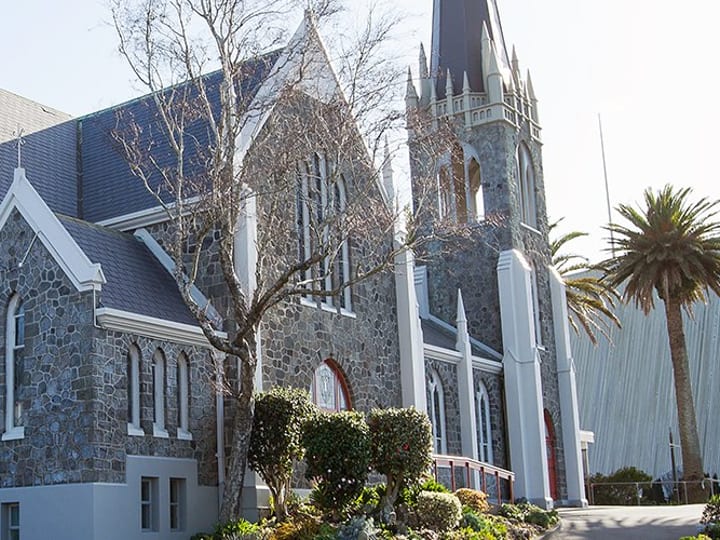 Large dark stone Gothic church with spire, multiple arched windows, and adjacent modern wing