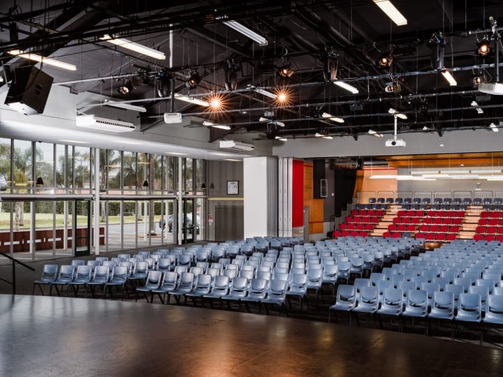 Multi-purpose hall with stage, tiered seating, and exposed black ceiling grid with stage lighting