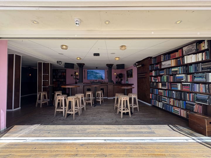 Pink-walled interior room with timber bar, bookshelves floor-to-ceiling, and backless stools on wooden floor