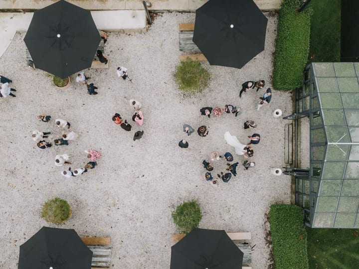 Square-cropped aerial of gravel courtyard with parasols and wedding guests