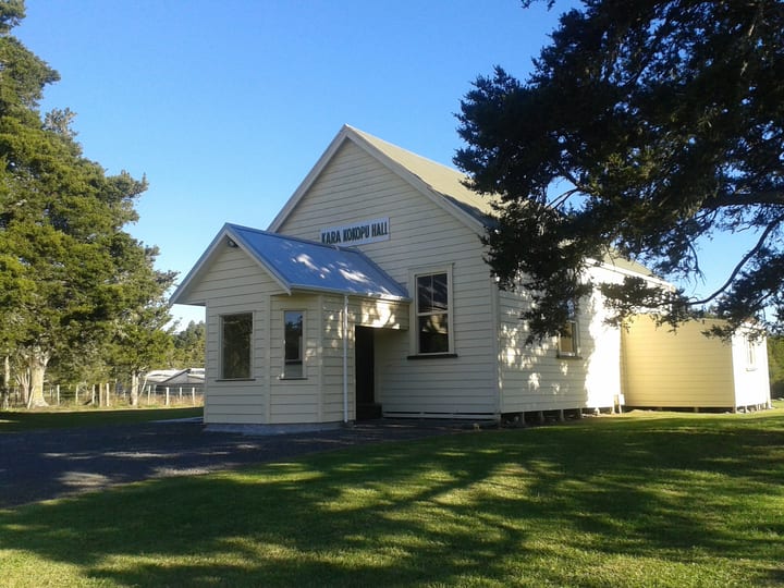 Small white timber hall with metal roof and signage on grassed section