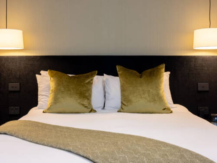 Hotel bed with dark bedhead and gold-green velvet cushions, warm bedside lighting