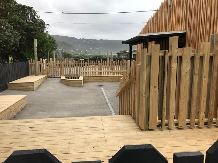 Timber-battened building exterior with open courtyard, low timber seating and fencing, overcast hillside backdrop