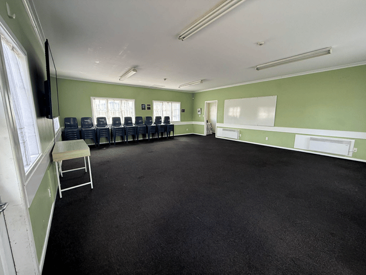 Plain hall with dark carpet, green walls, stacked chairs, and whiteboard