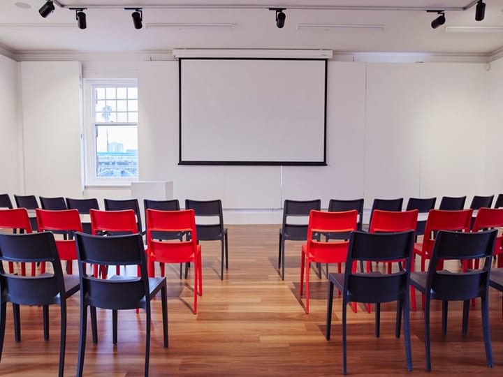 White-walled room with red and dark chairs in rows, projection screen, timber floor