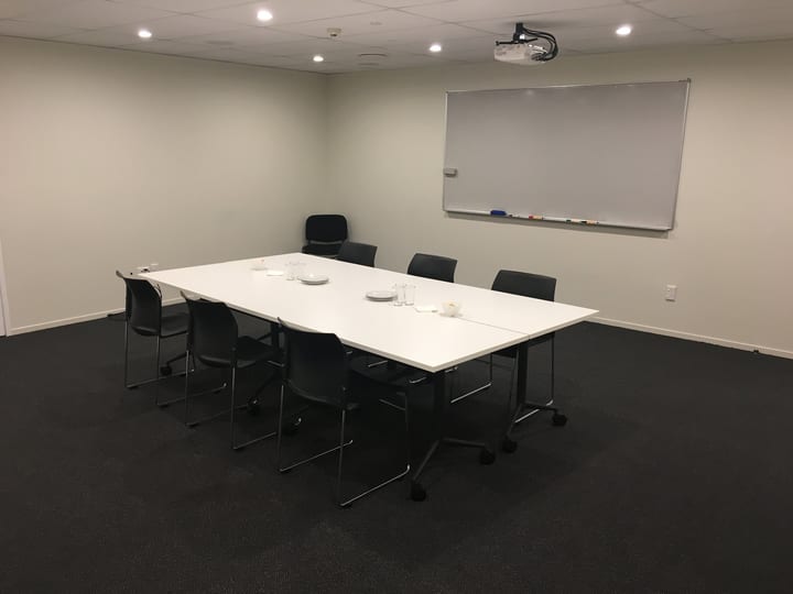 Plain meeting room with a white rectangular table, dark chairs, whiteboard, and projector