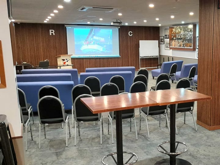 Timber-panelled function room with rows of blue and black chairs facing a projection screen and lectern