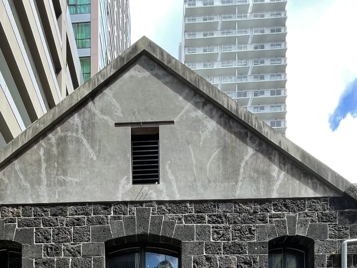 Dark basalt stone warehouse exterior with arched entry and gabled roofline, flanked by city towers