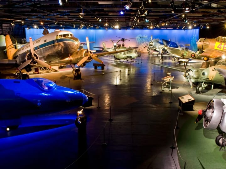 Large darkened museum hall with multiple historic aircraft on display under coloured spotlights