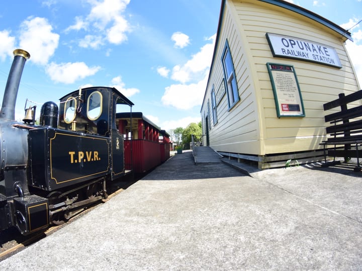 Historic steam locomotive and carriages alongside a station building labelled Opunake Railway Station