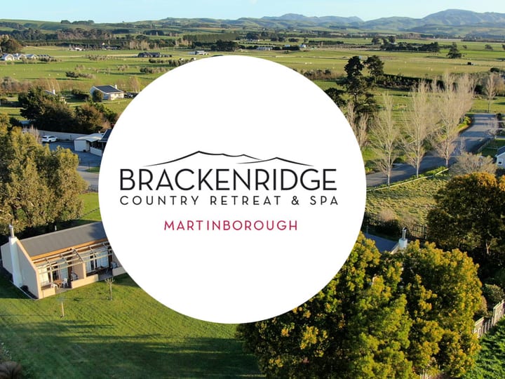 Aerial view of low-rise white buildings set among green farmland, rolling hills and flat plains extending behind, venue logo overlaid