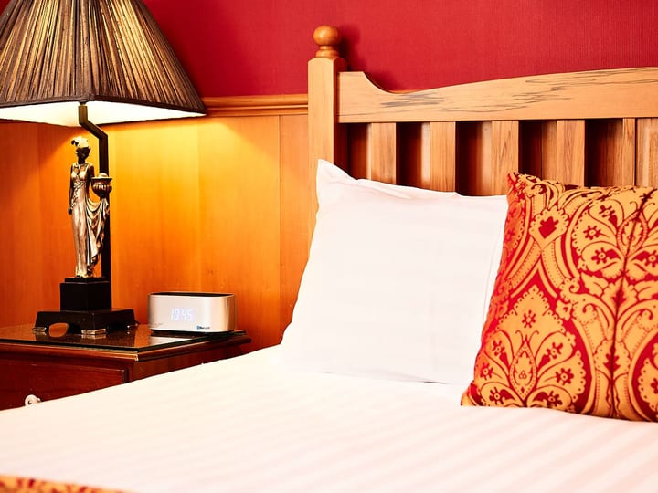 Hotel bedroom with timber headboard, red patterned cushion and bedside lamp, warm tones