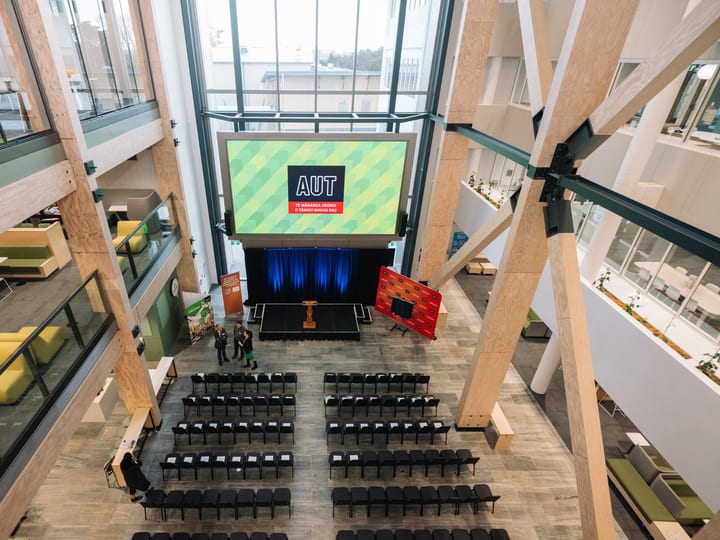Large atrium event space with timber columns, rows of chairs, and a raised screen displaying a logo
