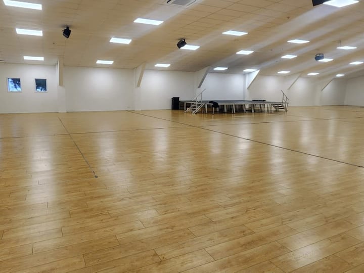 Large empty hall with timber floor and raised platform at the far end, lit by overhead panels