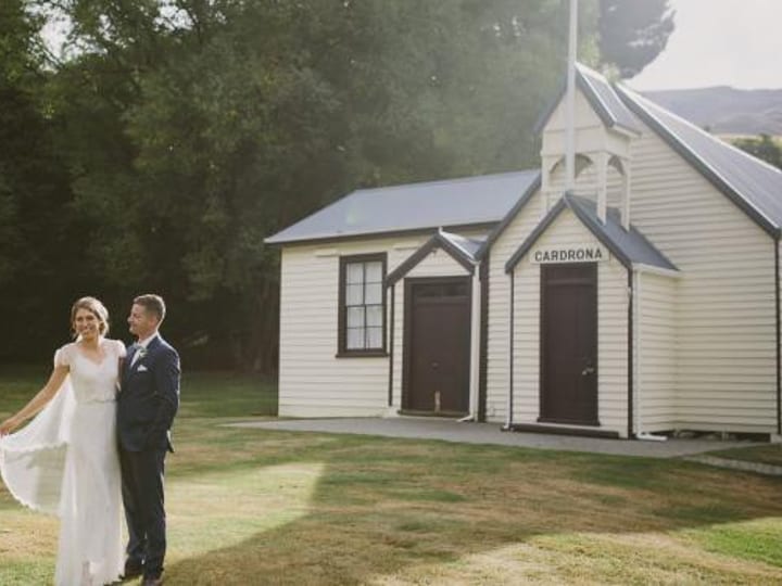 A couple in wedding attire on a lawn beside a small white weatherboard chapel, framed by tall trees with hills behind
