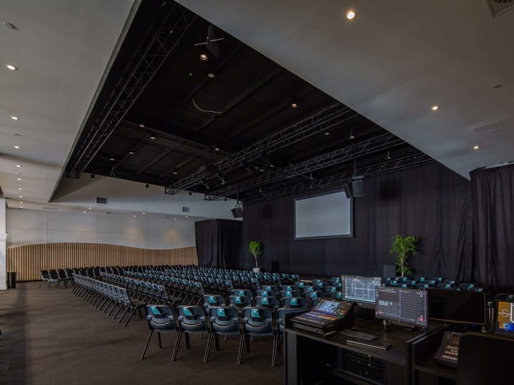 Conference hall with rows of chairs facing a projection screen, lighting truss overhead, and a production desk