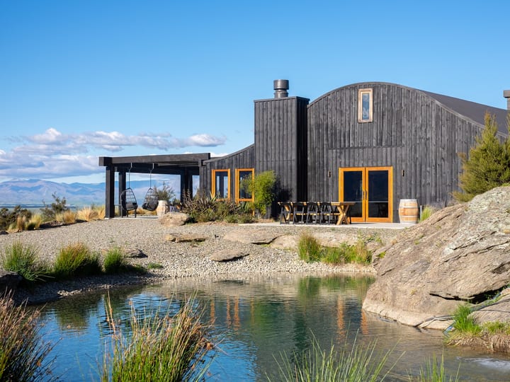 Dark timber barn with curved roof beside a pond, rocky landscape, open mountain backdrop