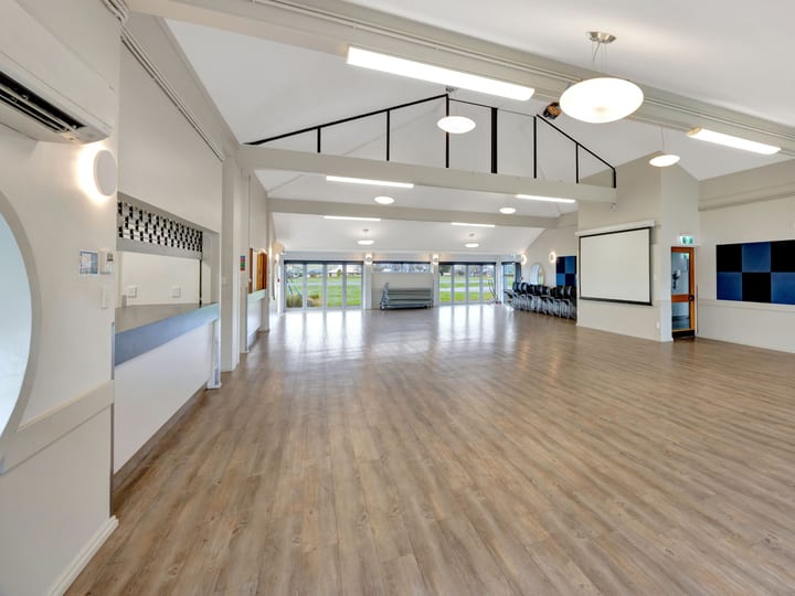 Empty community hall with timber floor, high ceiling, and projection screen