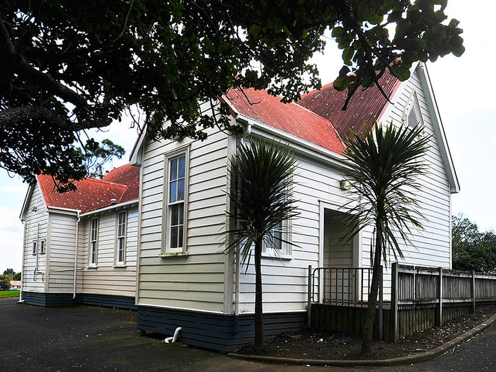 White weatherboard hall with red corrugated roof and cabbage trees at entrance
