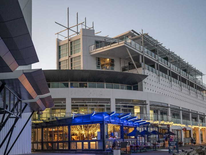 Waterfront venue pavilion with blue neon roof fins and glazed walls, dusk street view