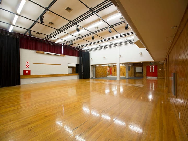 Large hall with polished timber floor, stage lighting rig, and heavy curtains